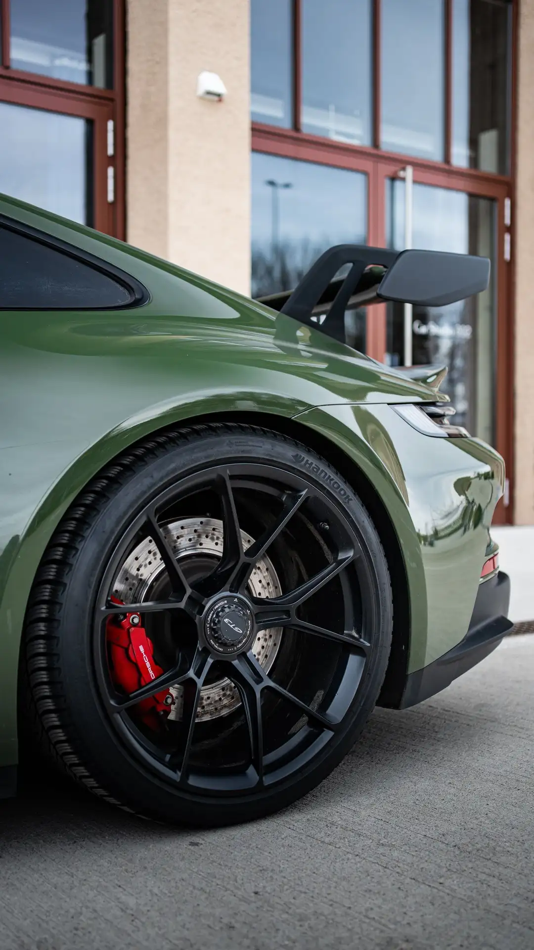 Close-up of a back wheel of our olive green Porsche GT3 in front of an industrial building. 